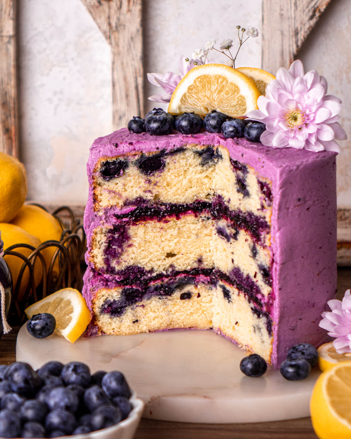 cut open blueberry lemon cake frosted with purple frosting, filled with blueberry jam, topped with blueberries, lemon slices and flowers