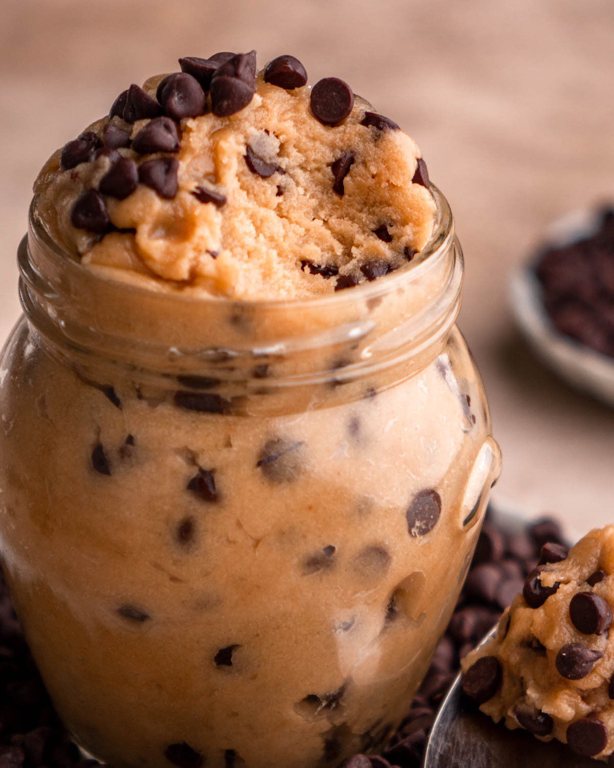 cookie dough in a small jar with a scoop taken out, on a plate of chocolate chips with a spoon of cookie dough next to it