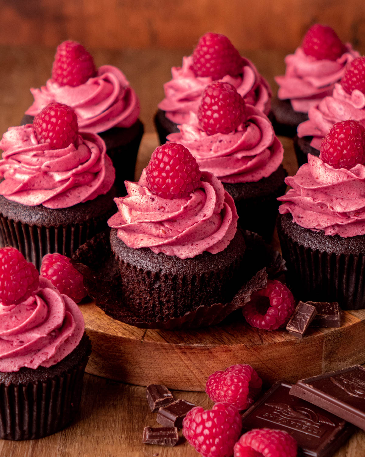 chocolate raspberry cupcakes on a wood platter with raspberries and chocolate around, the center cupcake with the wrapper taken off