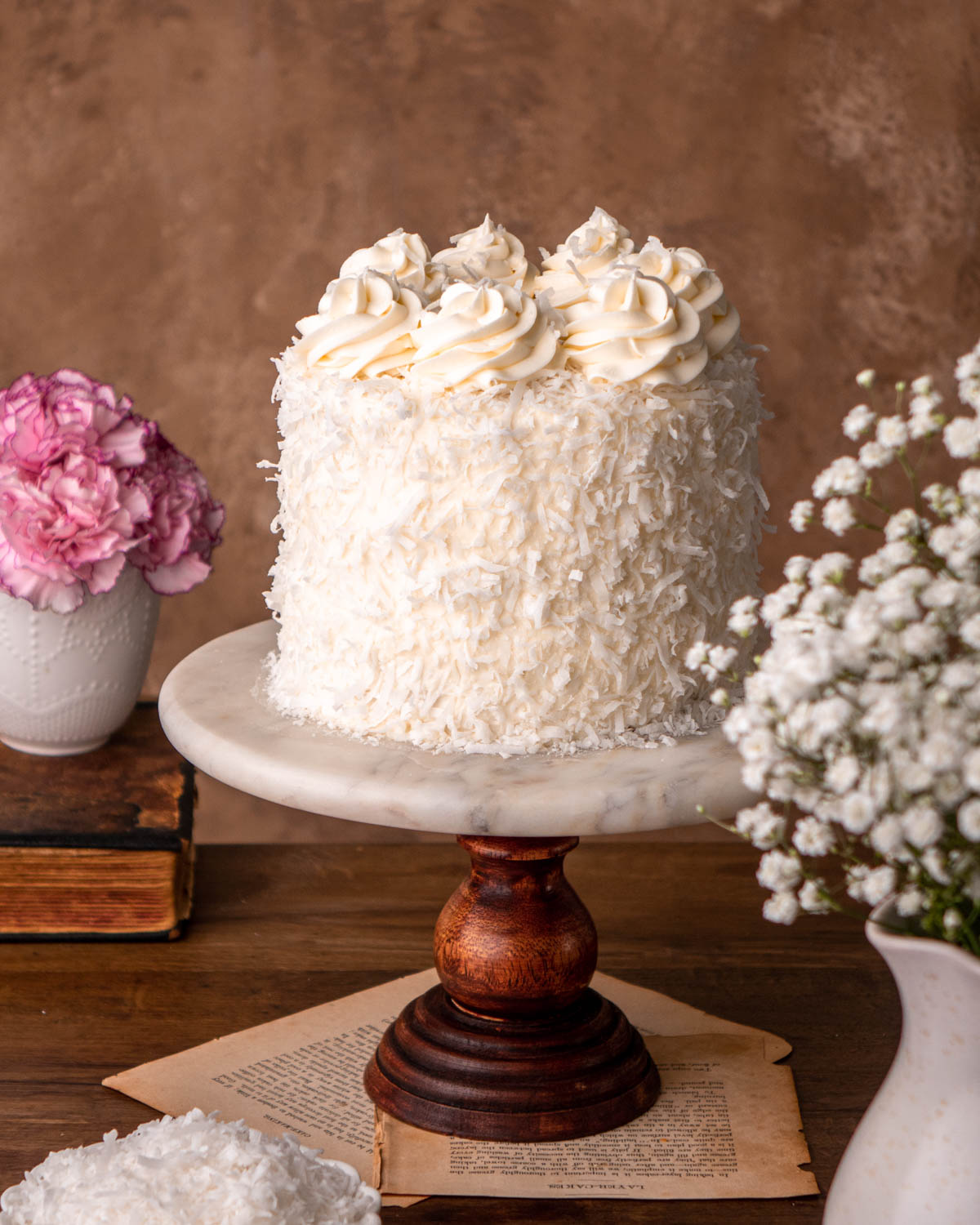 coconut cake with shredded coconut pressed on outside of the cake with swirls of cream cheese frosting on top, on a marble cake stand on a wood board
