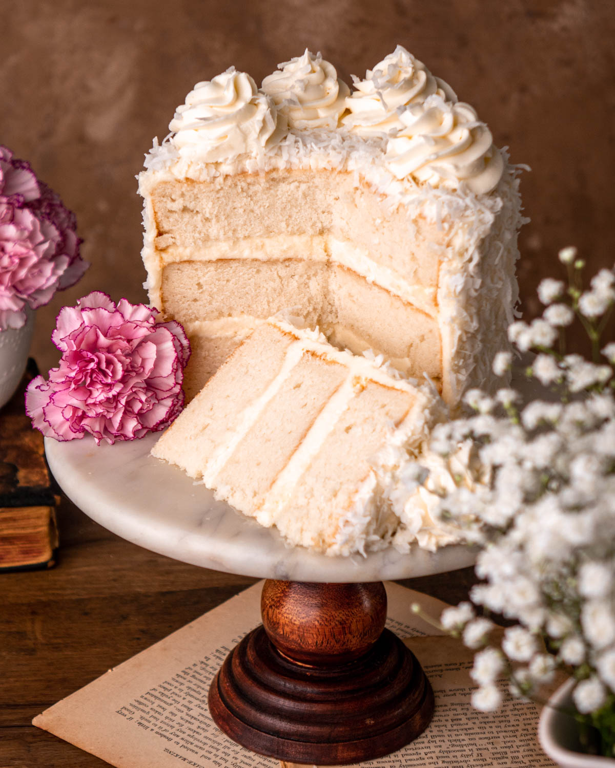coconut cake cut open, with cake slice in front, on a marble cake stand with pink and white flowers