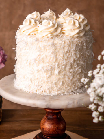 coconut cake with shredded coconut pressed on outside of the cake with swirls of cream cheese frosting on top, on a marble cake stand on a wood board
