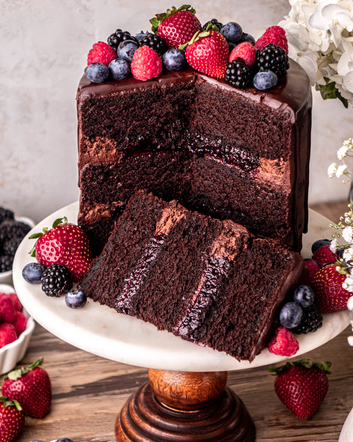 chocolate cake filled with berry jam cut open on a marble and wood cake stand, cake slice in front of it, topped with strawberries, blueberries, raspberries and blackberries 