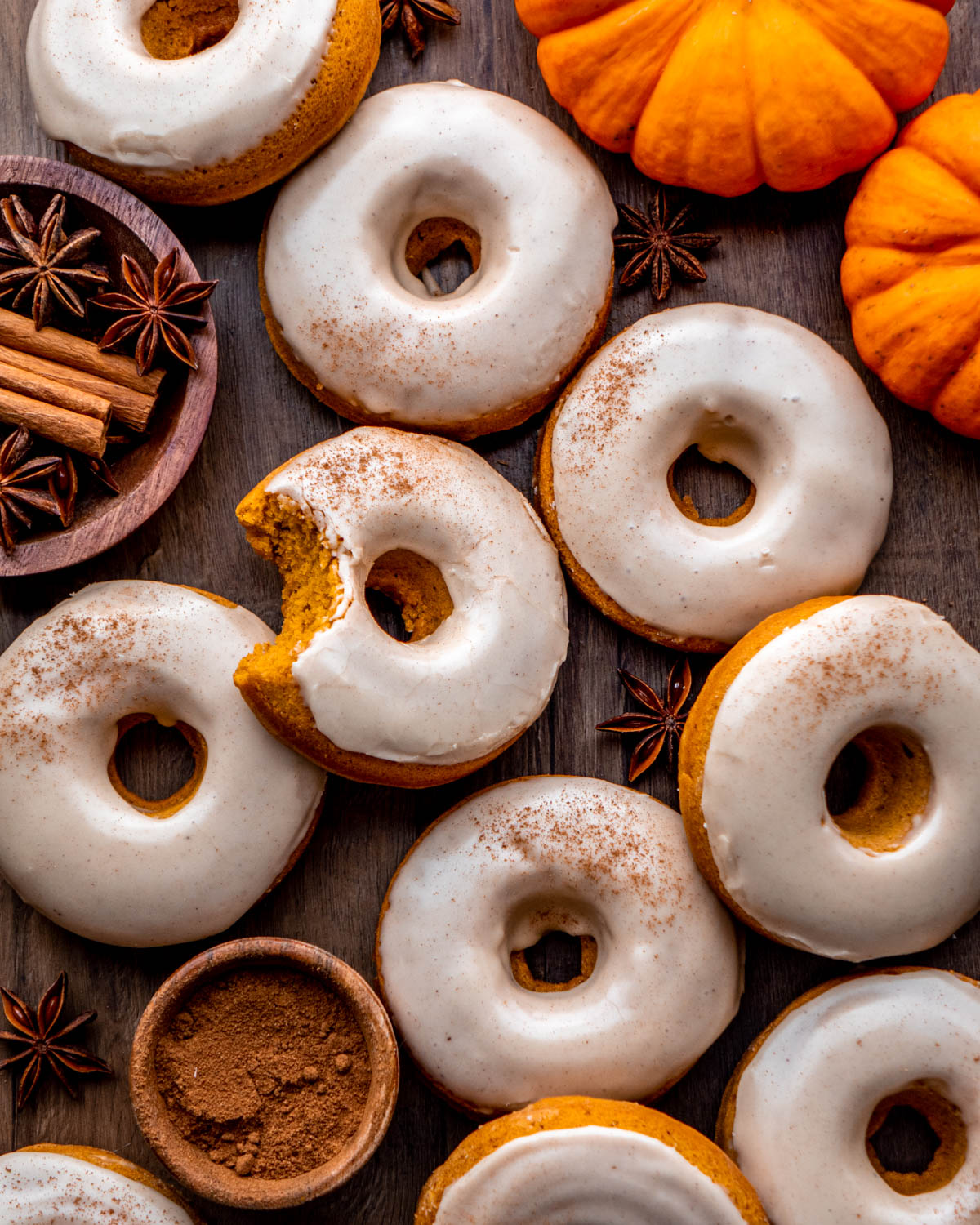 pumpkin donuts with a bite taken out on a wood board with pumpkins, cinnamon sticks and star anise around