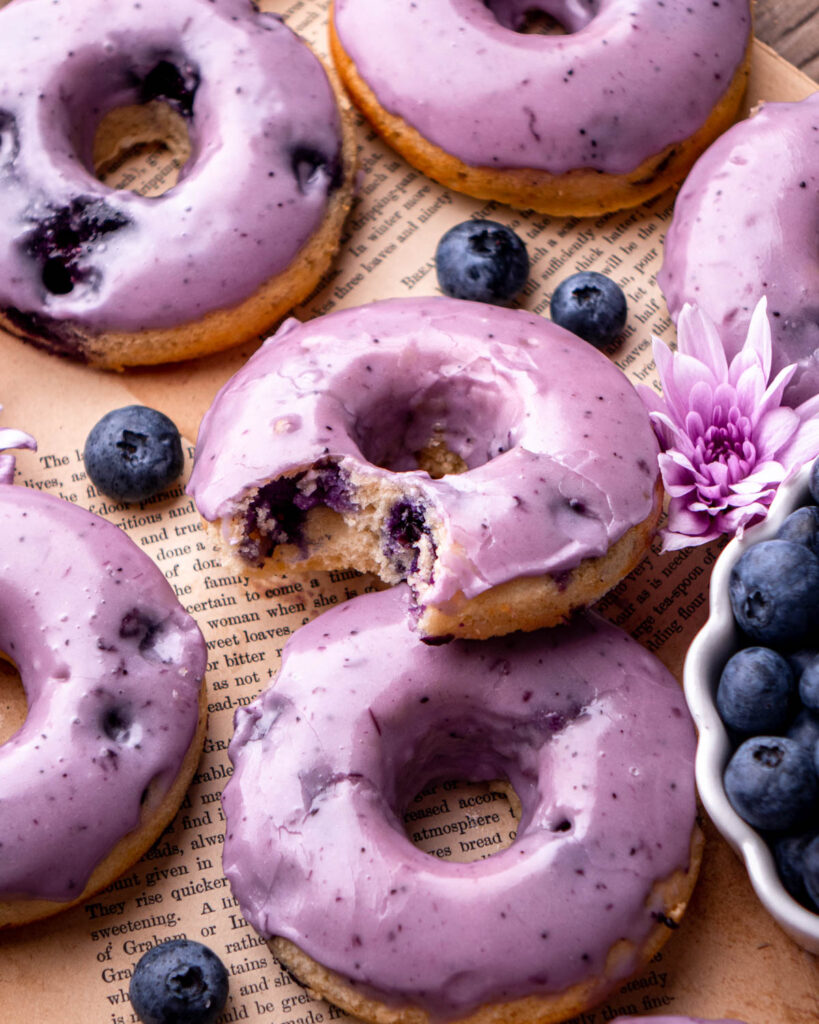blueberry donuts laid on top of old book pages, one with a bite taken out, with bowl of blueberries and flowers