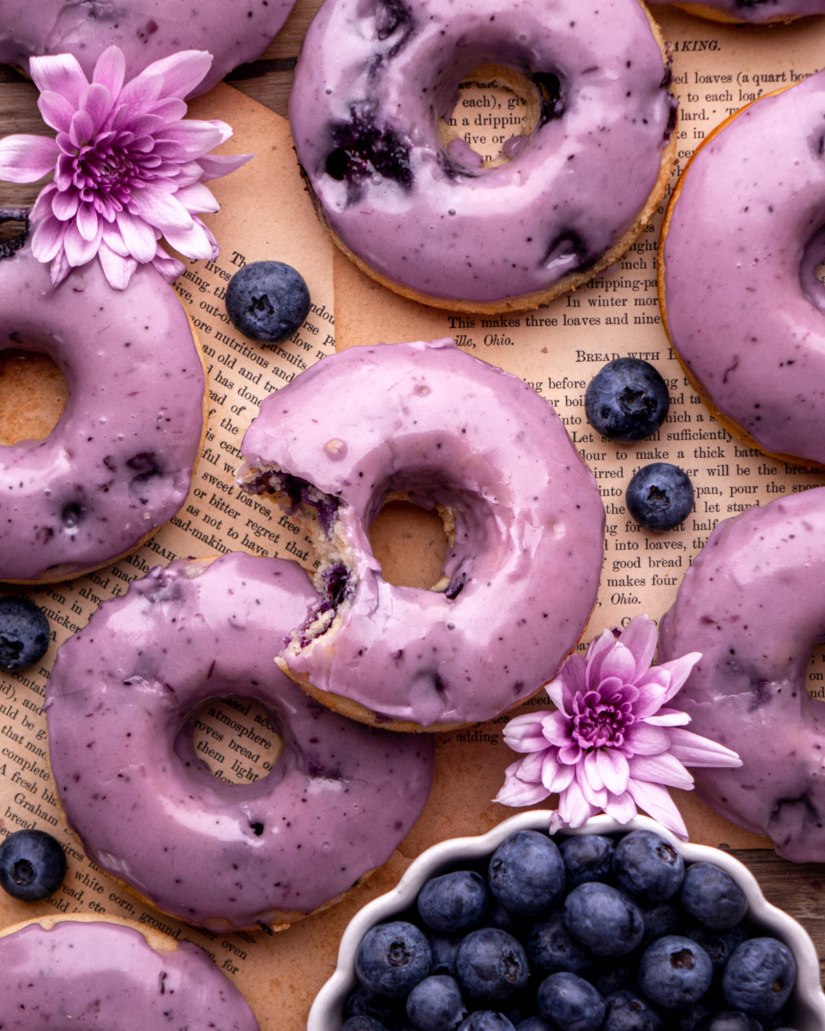 blueberry donuts laid on top of old book pages, one with a bite taken out, with bowl of blueberries and flowers