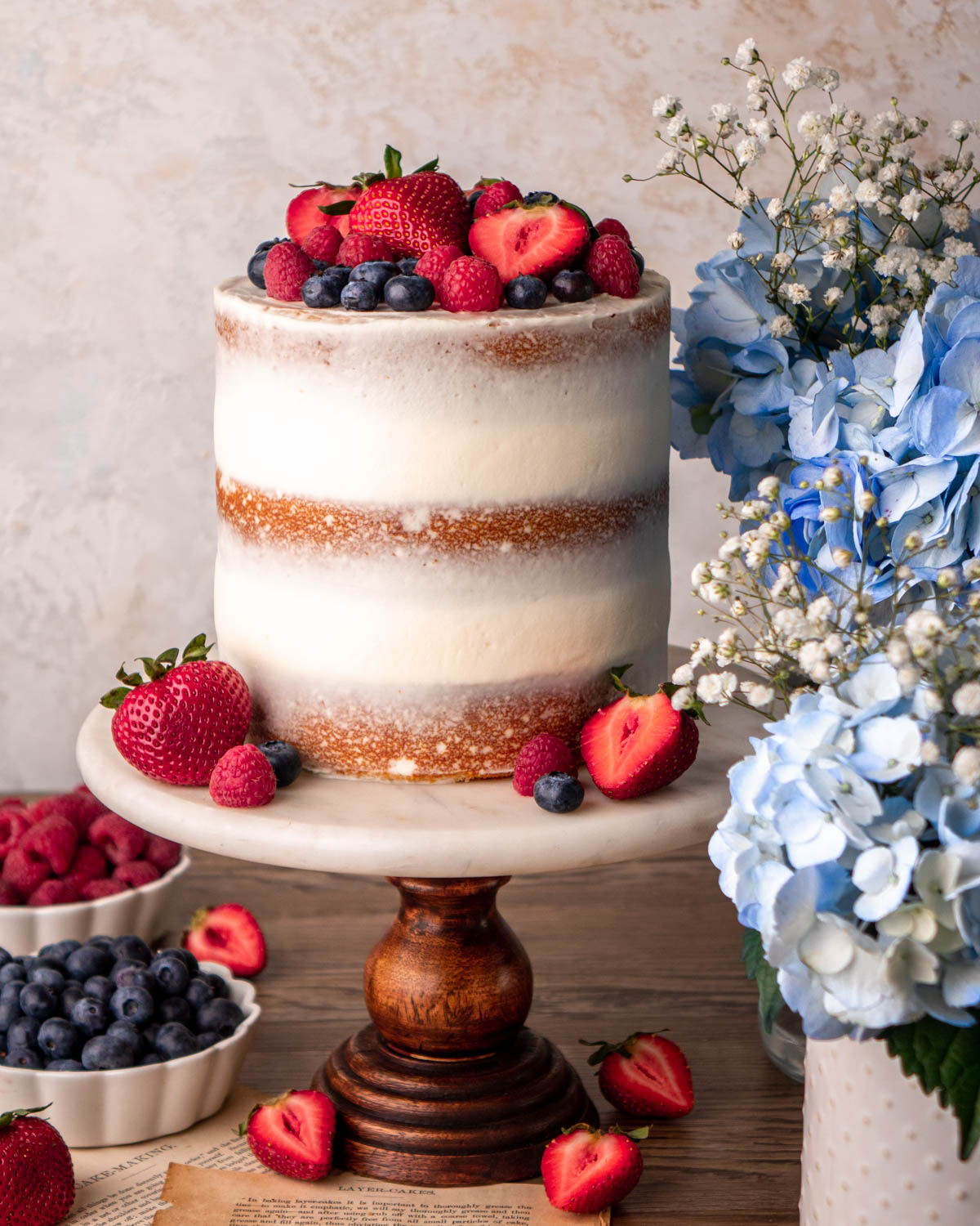 semi-naked Chantilly cake with blueberries, strawberries and raspberries on top, on a marble cake stand on a wood board with blue flowers around it
