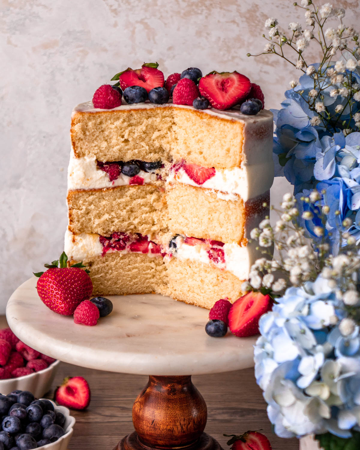 cut open Chantilly cake with blueberries, strawberries and raspberries on top, on a marble cake stand on a wood board with blue flowers around it
