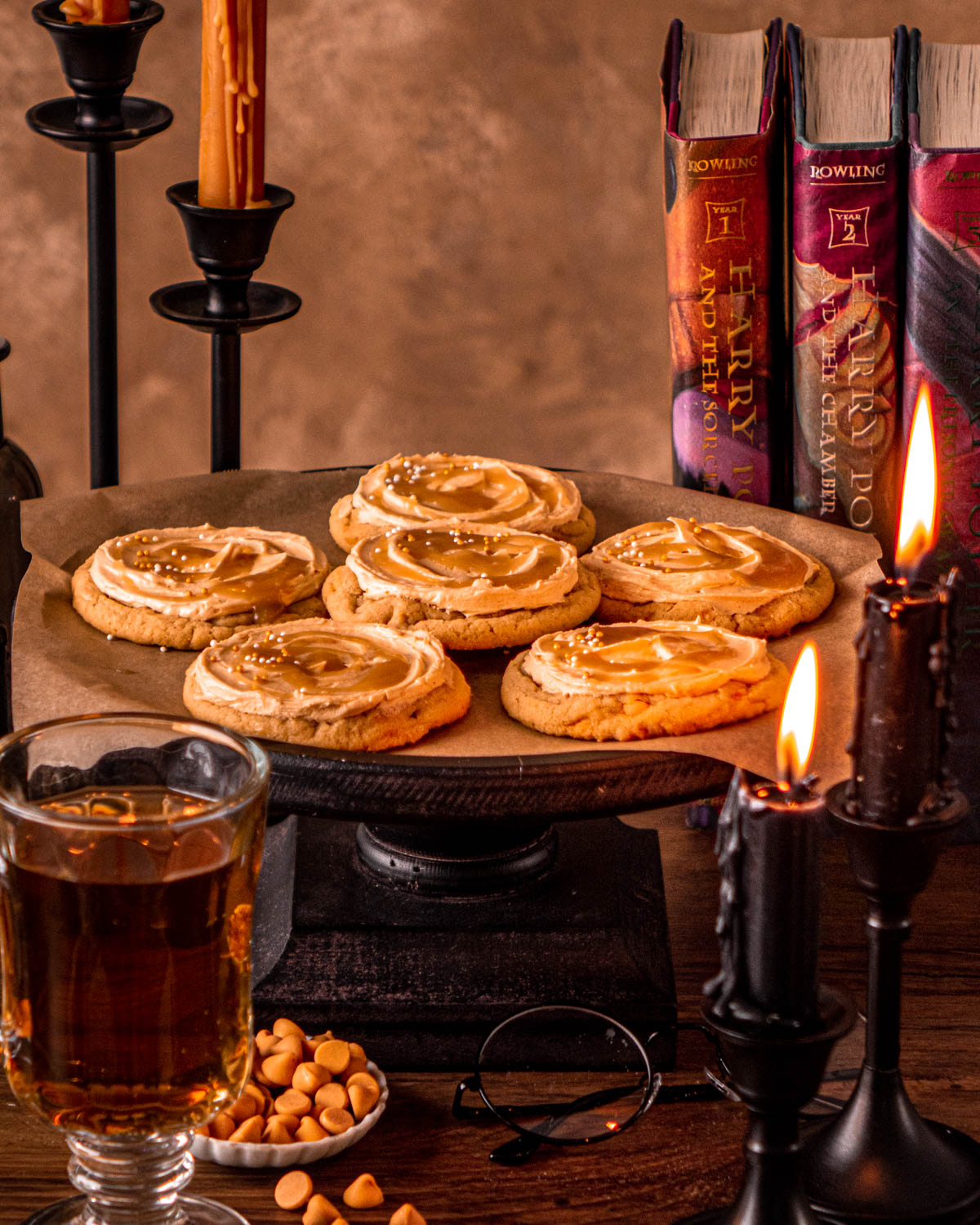 butterbeer cookies on a cake stand with candles, books and drinking glass around