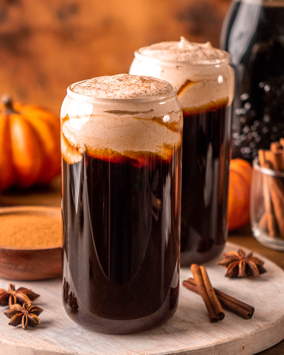  two glasses of cold brew coffee topped with pumpkin cold foam, pumpkins, a jar of coffee and cinnamon sticks in the background