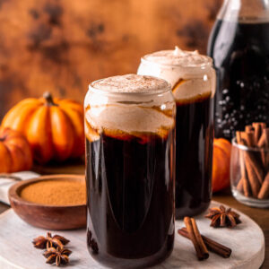 two glasses of cold brew coffee topped with pumpkin cold foam, pumpkins, a jar of coffee and cinnamon sticks in the background
