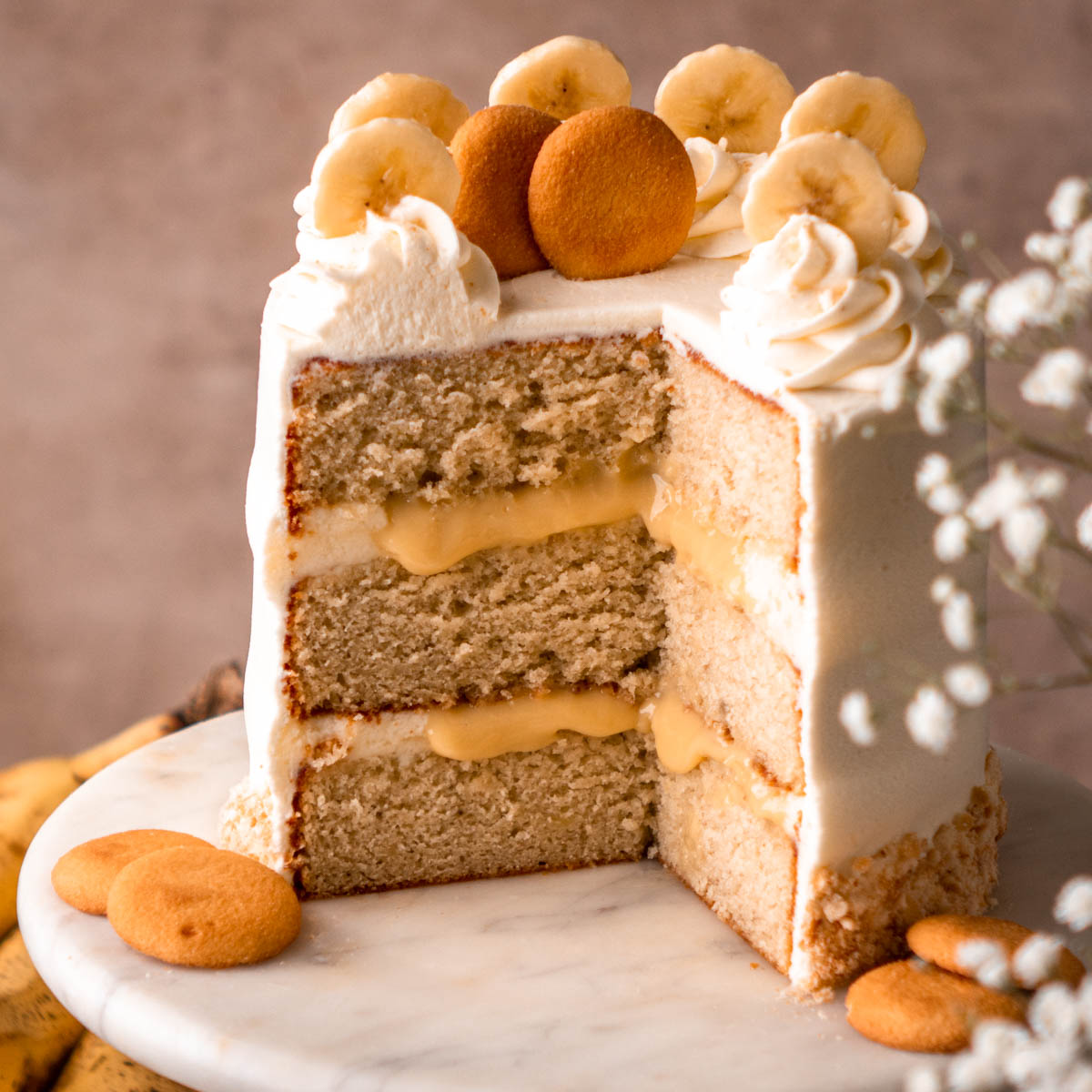 banana cream cake decorated with banana slices and Nilla Wafers, cut open with banana pudding filling, on a white marble cake stand with bananas in the background