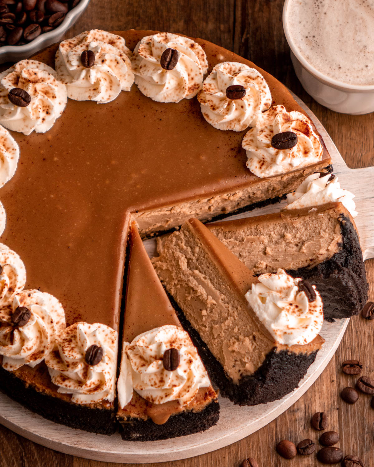 Coffee cheesecake with three slices laying on each other, on a white wooden serving plate with a cup of coffee and coffee beans around