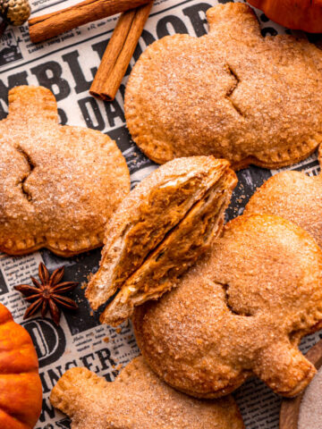 pumpkin pasties arranged on newspaper with pumpkins around, one cut in half in the center so the filling is visible