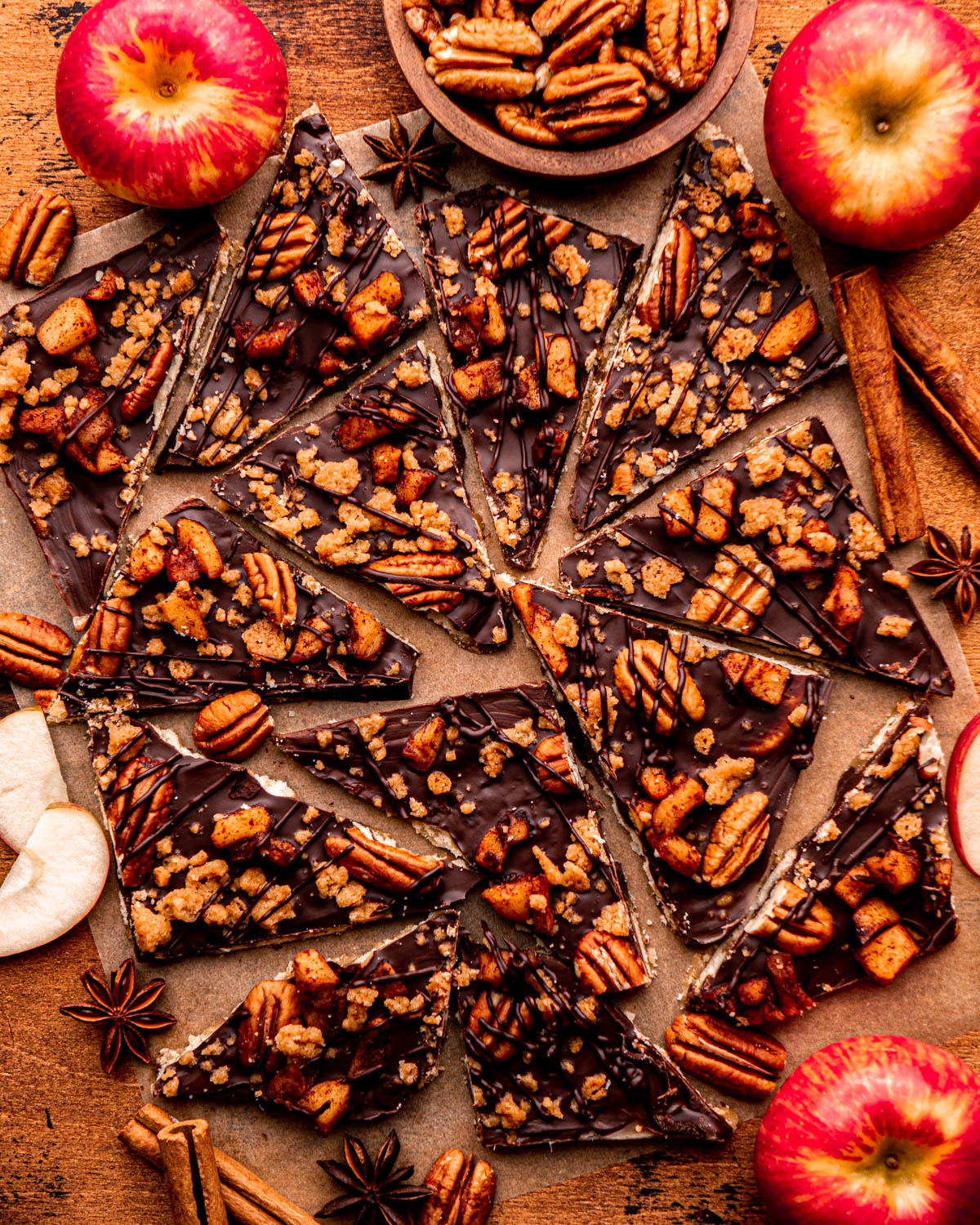 Thanksgiving cracker toffee pieces arranged on parchment paper with cinnamon sticks, apples and pecans near by