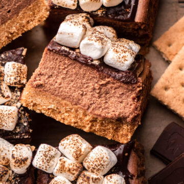 close up of a s'mores cheesecake bar arranged on parchment paper with other cheesecake bars, graham crackers and chocolate around